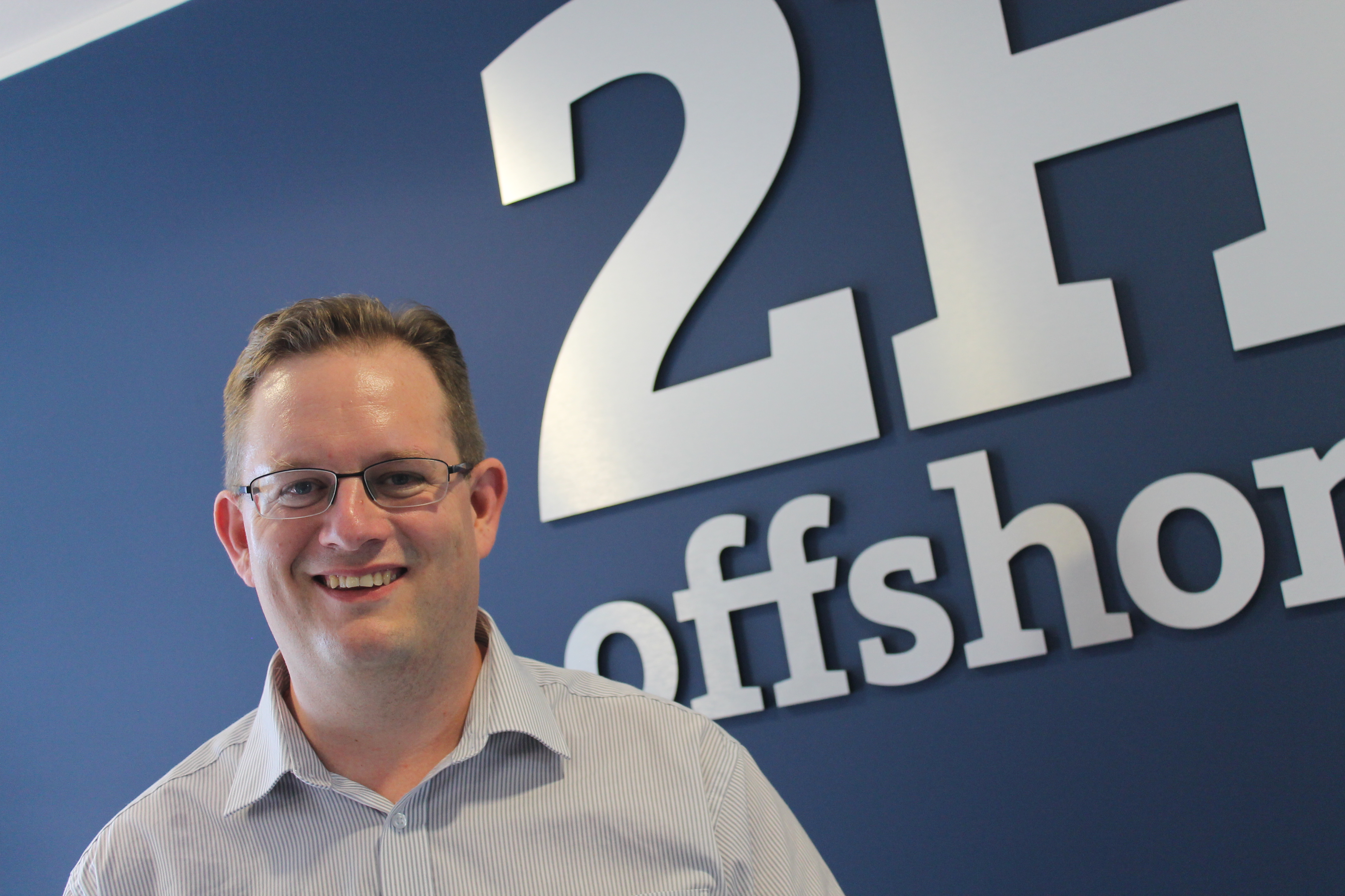 Man wearing glasses and shirt smiling at the camera, with a logo in the background